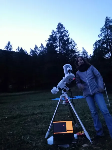 Crépuscule. Une jeune femme pose à côté des téléscopes. Un ordinateur portable est déployé sous le télescope.